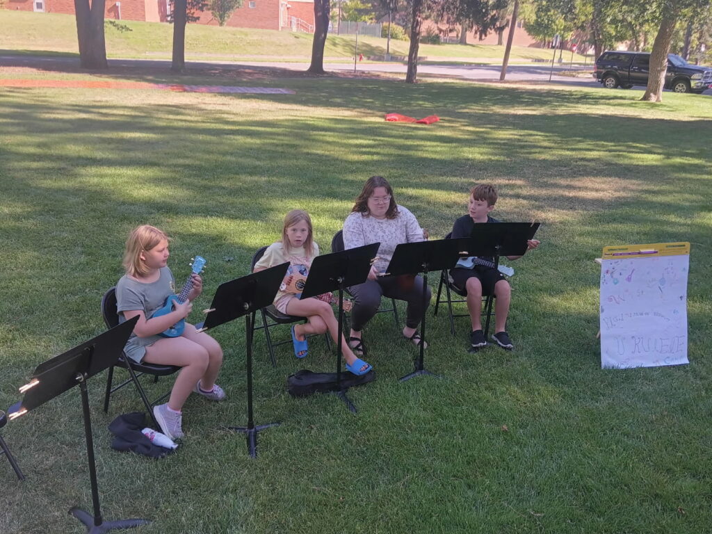 Three children and their instructor performing ukulele songs in the park.